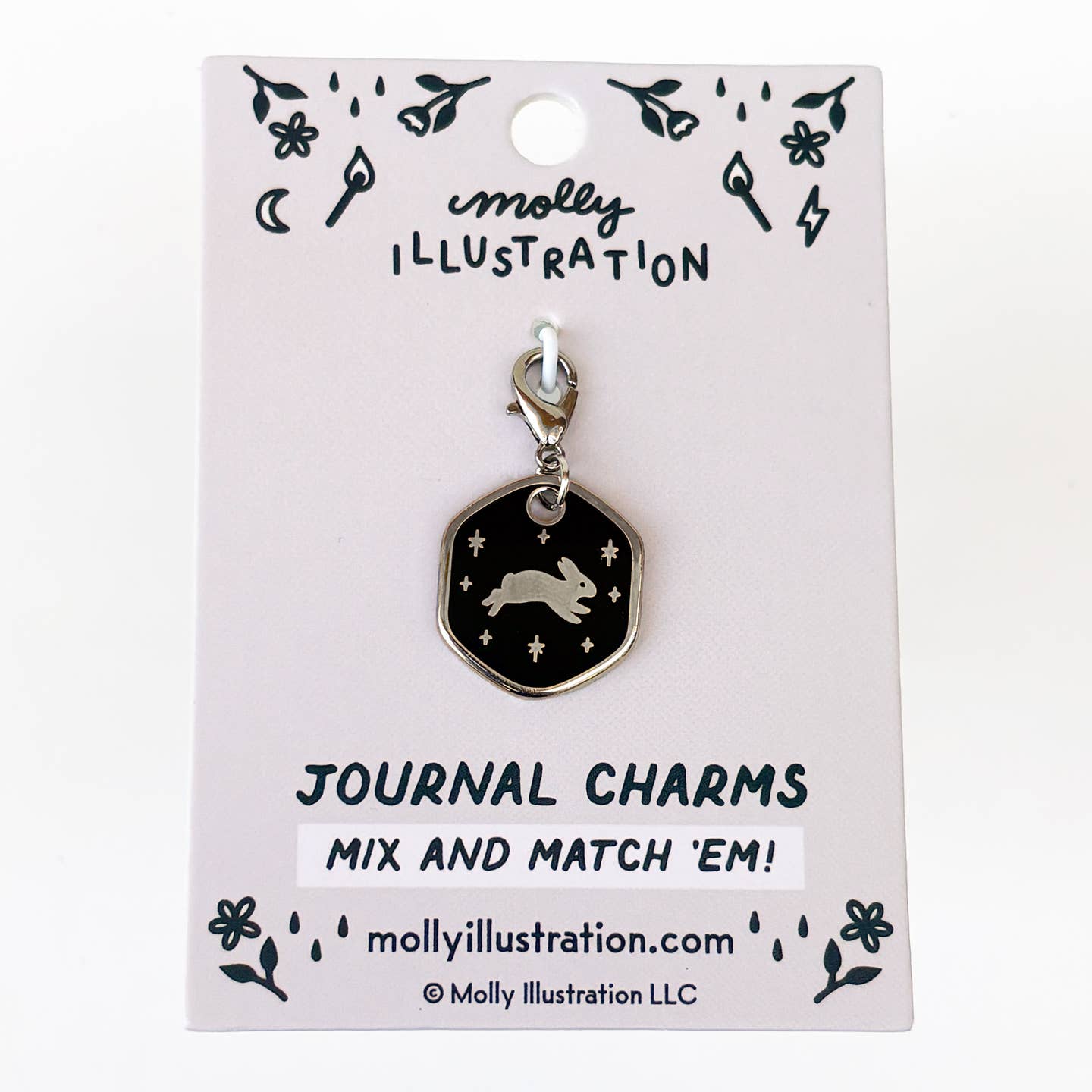 A silver-toned journal charm by Molly Illustration, featuring a black hexagonal pendant with a silver rabbit and stars, displayed on its light pink backing card.