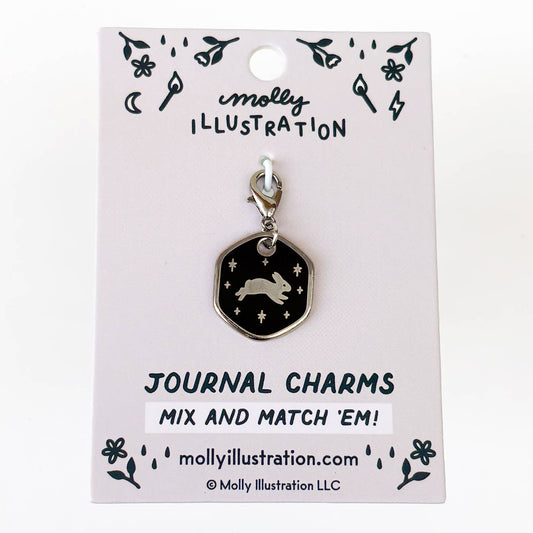 A silver-toned journal charm by Molly Illustration, featuring a black hexagonal pendant with a silver rabbit and stars, displayed on its light pink backing card.