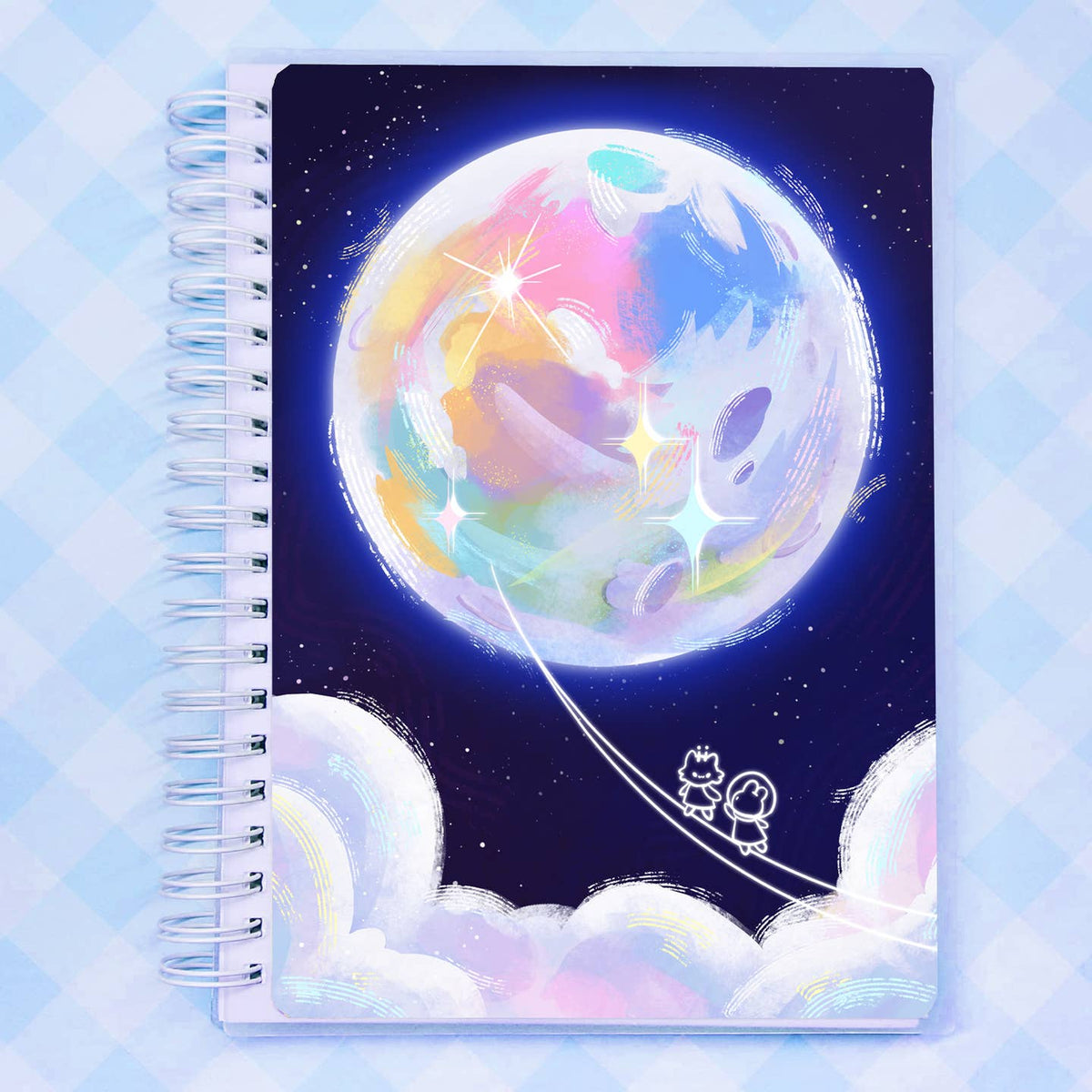 A spiral-bound reusable sticker notebook with a colorful pastel illustration of a glowing moon, stars, and two small cartoon figures, a cat and bunny, on a dark blue night sky, set against a light blue checkered background.