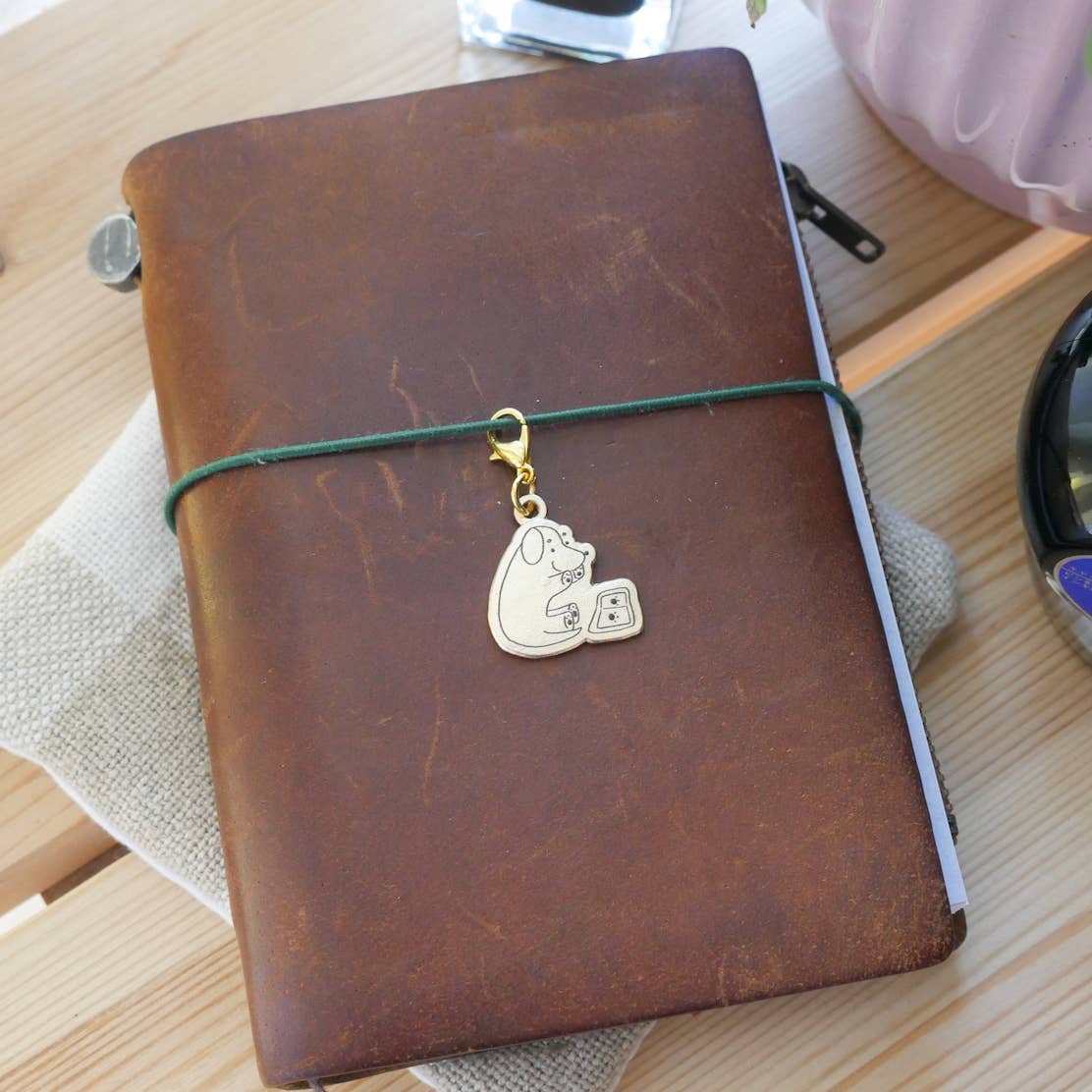 Small brass "Paw Prints" charm engraved with an illustration of a cute dog stamping their paw prints in a journal with a gold-tone clasp attached to a brown leather Traveler's style jounral.