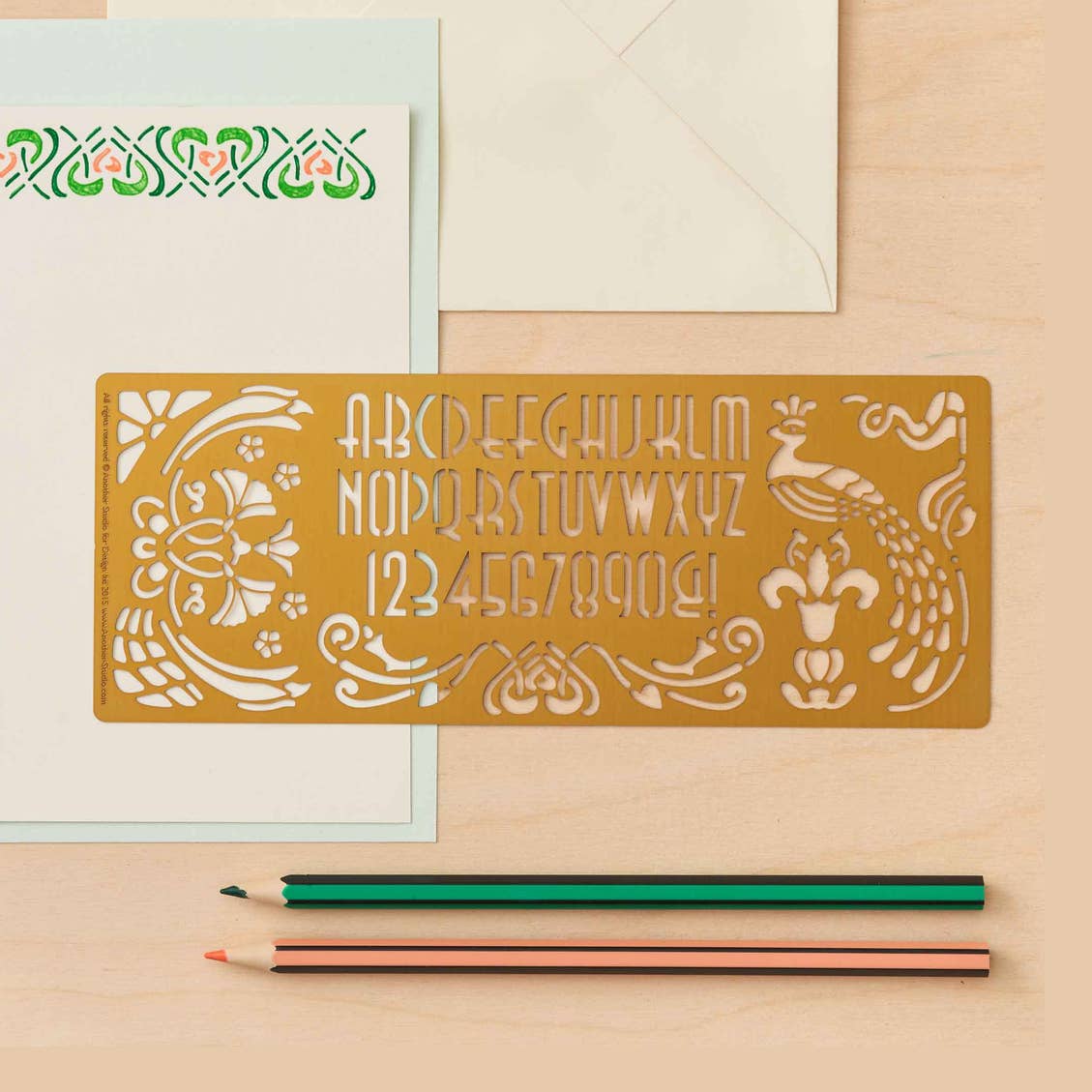 Decorative brass Art Nouveau ruler with letter and number designs on a peach surface with a pencil and paper.