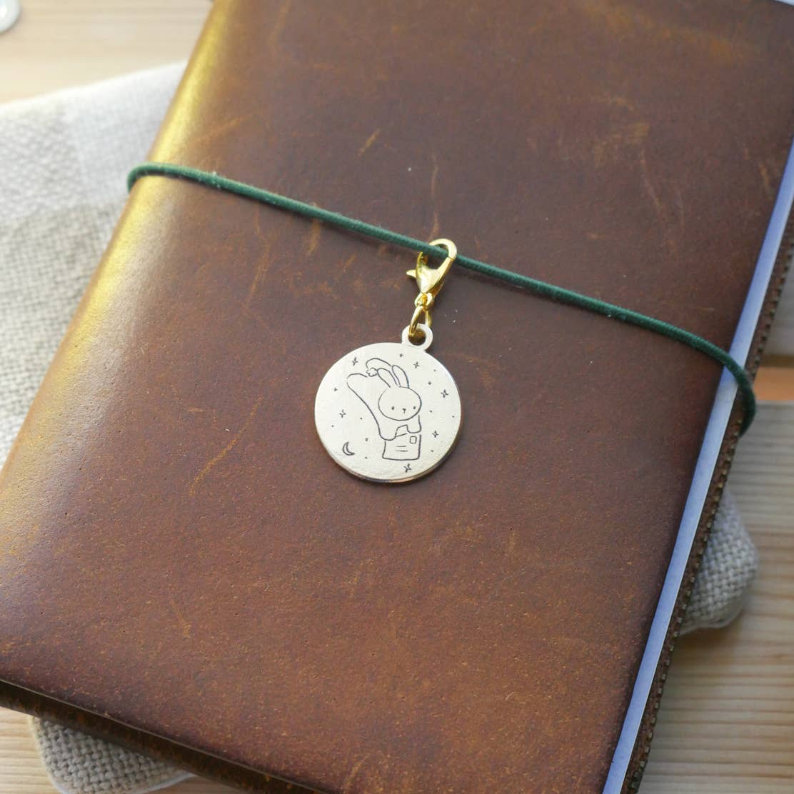 Small brass "For You" charm engraved with an illustration of a leaping bunny, holding an envelope, with stars and moon in the background, with a gold-tone clasp attached to a brown leather Traveler's style jounral.