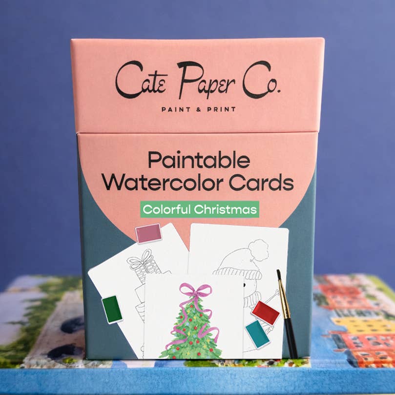 A boxed set of Cate Paper Co. Paintable Watercolor Cards titled 'Colorful Christmas,' shown standing upright on a blue surface against a blue background.