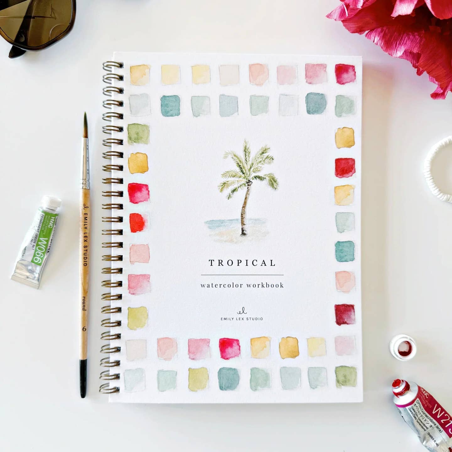 The front cover of the spiral bound Tropical watercolor workbook with a watercolor painted image of a palm tree surrounded by color swatches and a paintbrush and paint tube on a white surface
