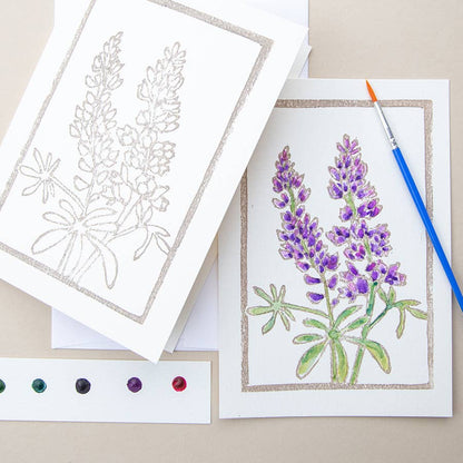 Card with an illustration of lupine flowers with watercolors and a paintbrush on a cream background